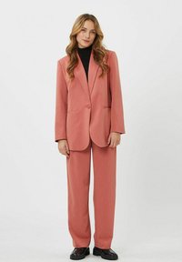 Conjunto de blazer y pantalones en rosa empolvado con un diseño estructurado, que presenta una tela suave y cierre de un solo botón. Un cuello alto negro por debajo.