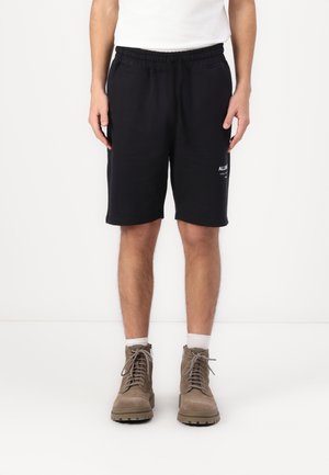 Pantaloncini neri in cotone con fascia elastica in vita e tasche laterali, con un piccolo logo bianco. Indossati con stivaletti marroni stringati e calzini bianchi.