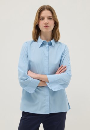 Junge Frau mit schulterlangen blonden Haaren, die ein hellblaues Hemd mit Knöpfen und dunkle Hosen trägt, steht mit verschränkten Armen.