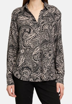 Femme portant une chemise à manches longues noire et beige à boutons avec un motif abstrait de fleurs et de feuilles, associée à un pantalon noir.