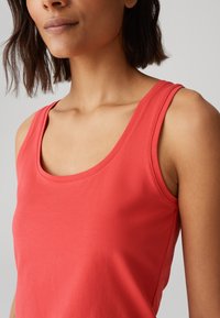 Top in corallo con scollo a barca, realizzato in un tessuto morbido e elasticizzato. Presenta spalline larghe e una texture liscia.