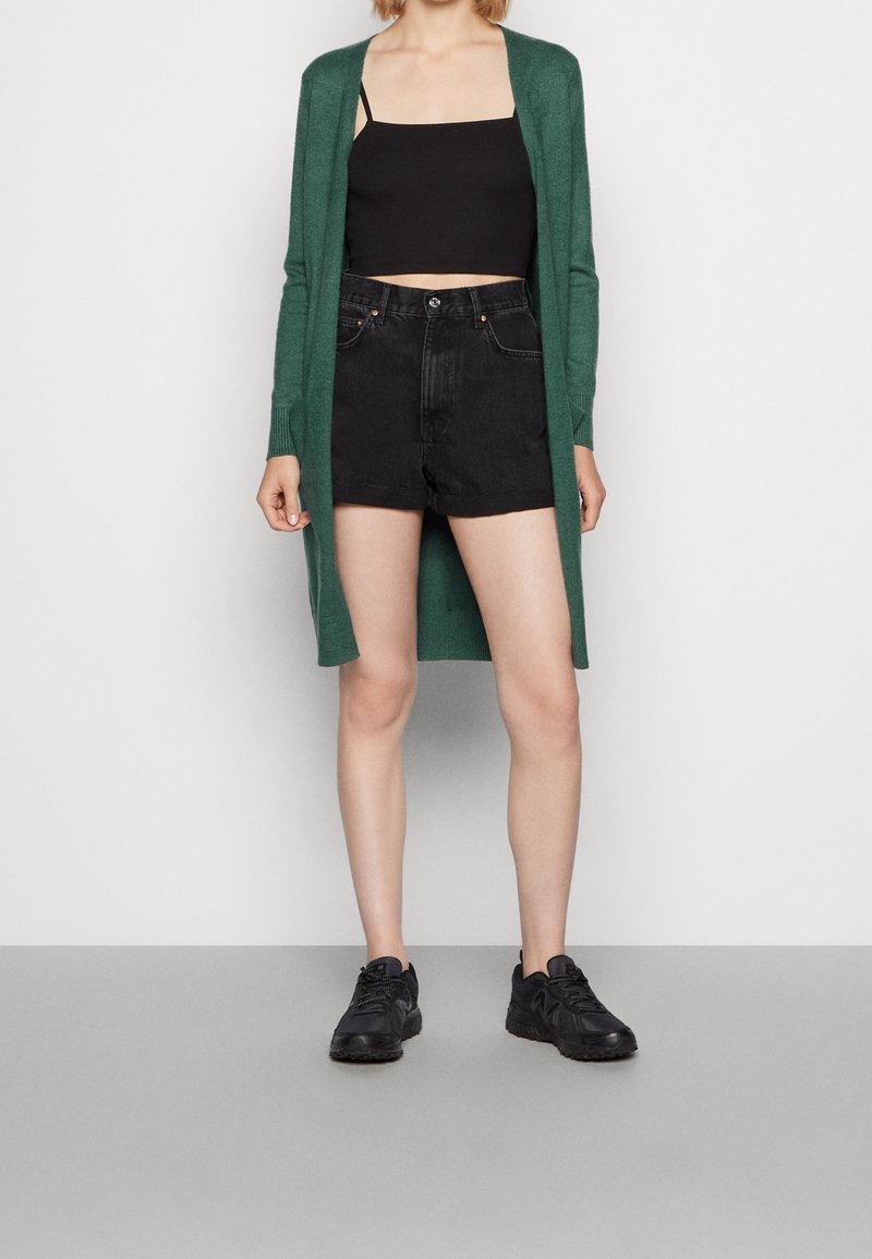 Personne portant un long cardigan vert, un crop top noir, un short taille haute noir et des baskets noires, debout sur un sol gris.