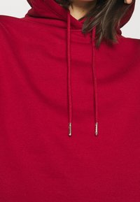 Röd huvtröja med en slät textur, med två dragsnören med metallspetsar. Enfärgad, minimalistisk design, inga mönster.