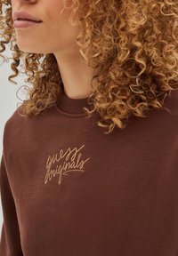 Sweatshirt marron avec col rond, présentant le texte brodé "guess originals" en or. Fabriqué en tissu doux et texturé avec une coupe décontractée.