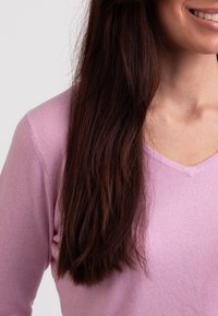 Rosa V-ringad tröja gjord av mjukt stickat tyg, med ribbat mönster och åtsittande design. Håret är långt och mörkt, fallande åt sidan.