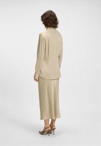 Woman with curly hair stands facing away, wearing a beige long-sleeve blouse, matching midi skirt, and taupe slingback heels.