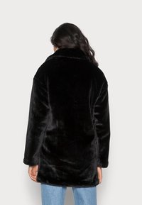 Abrigo de piel sintética negra con un cuello alto, con una textura suave y un corte oversize, visto desde atrás. Diseño minimalista, sin herrajes visibles.