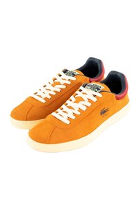 Sneakers in suede arancione con lacci bianchi; presentano un interno imbottito nero e un tallone rosso a contrasto. Logo Lacoste in blu navy.
