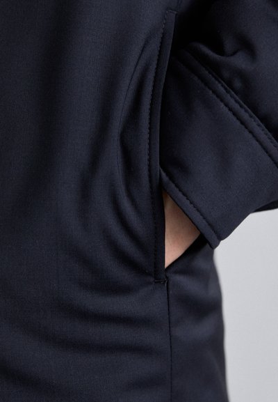 Indumento blu navy con tasca laterale e tessuto testurizzato. Il design include un dettaglio di cucitura visibile sulla manica.