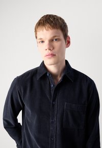Joven con cabello corto castaño claro y ojos azules, vistiendo una camisa de terciopelo de color azul marino oscuro, posando frente a un fondo claro y liso.