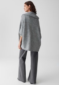 Grauer gestrickter Poncho mit weitem Kragen, überschnittenen Schultern und strukturiertem Finish. Kombiniert mit grauen, weit ausgestellten Hosen. Einfaches Design, gemütliche Passform.