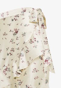 Haut enveloppant floral en tissu cream ; présente un bord à volants, des liens aux épaules, avec de petits motifs floraux roses et verts tout au long.