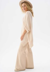 Beżowy dziany ponczo z szerokimi rękawami i bocznym rozcięciem, w połączeniu z jasnobeżowymi spodniami w stylu dzwonów i kapeluszem z szerokim rondem.