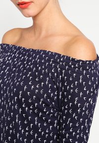 Topp navy av typen off-the-shoulder laget av bomull med et hvitt fuglemønster. Har elastiske rynker ved halslinjen og ermene.