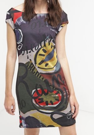 Kobieta w kolorowej sukience w abstrakcyjny wzór z żółtymi, czerwonymi, zielonymi i czarnymi kształtami oraz mazanym tekstem, stojąca na jednolitym tle.