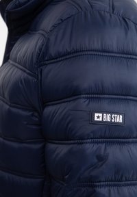 Tamsiai mėlynas pūkinis švarkas su siuvinėtu raštu, turintis ryškų „BIG STAR“ etiketę ant kairio peties ir lygų, blizgantį paviršių.