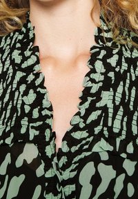 Mustapaita vihreällä abstraktilla kuviolla, jossa on röyhelökaulus ja teksturoitu kangas, esitellen kerroksellista muotoilua ja virtaavaa ilmettä.