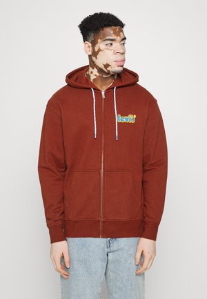 Sudadera con cremallera marrón, con cordones y bolsillos frontales; presenta un colorido logo bordado en el lado izquierdo del pecho. Lleva puesta sobre unos jeans de color azul claro.
