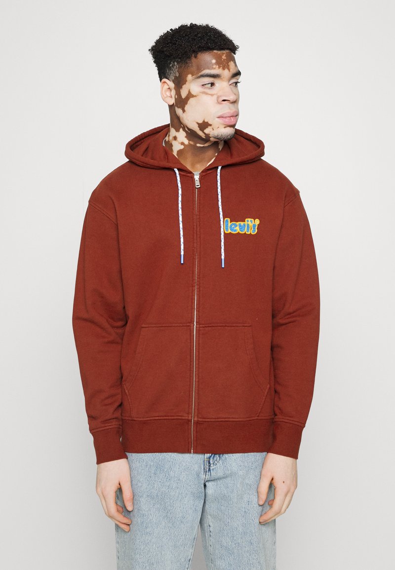 Sudadera con cremallera marrón, con cordones y bolsillos frontales; presenta un colorido logo bordado en el lado izquierdo del pecho. Lleva puesta sobre unos jeans de color azul claro.