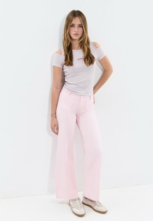 Jeune femme debout contre un mur blanc, portant un haut rayé à épaules dénudées, un pantalon large rose clair et des baskets beiges.