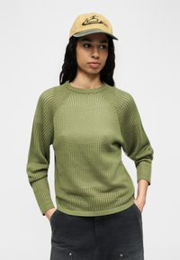 Νεαρή γυναίκα που φοράει ανοιχτό κίτρινο καπέλο, πράσινο πουλόβερ ανοιχτής πλέξης και σκούρο παντελόνι, με τα χέρια στις τσέπες, κοιτάζοντας ελαφρώς προς τα δεξιά.