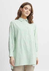 Camicia a righe verde e bianca con maniche lunghe, tasca sul petto e un orlo posteriore leggermente più lungo. Realizzata in tessuto leggero.