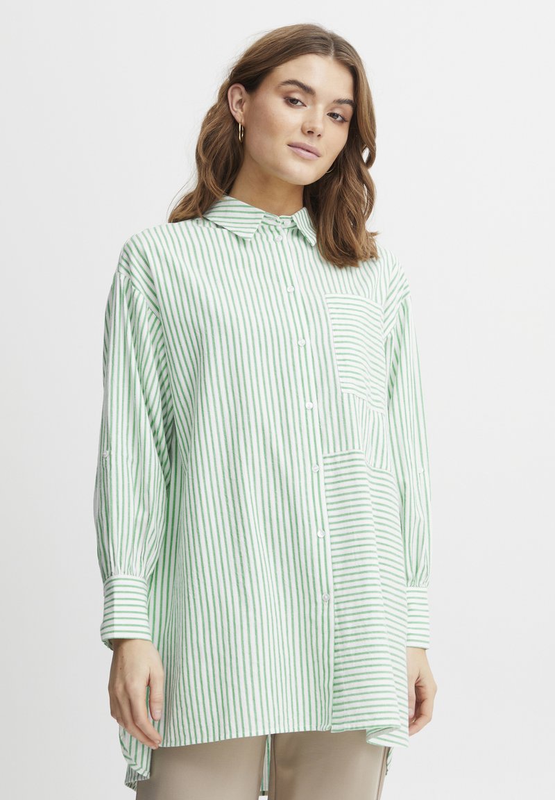 Camicia a righe verde e bianca con maniche lunghe, tasca sul petto e un orlo posteriore leggermente più lungo. Realizzata in tessuto leggero.