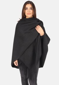 Schwarzer Wollponcho mit fransigen Kanten, der über eine Schulter drapiert ist. Glatte Textur, oversized Design, getragen von einer Person vor einem weißen Hintergrund.