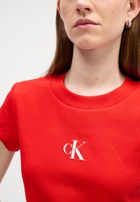Červené ribované tričko s kulatým výstřihem a krátkými rukávy, na přední straně s bílým logem "CK". Hladká textura a přiléhavý střih.