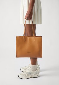 Borsa a spalla in pelle marrone chiaro con design minimalista, struttura piatta e due manici. Presenta un piccolo logo impresso sul davanti.