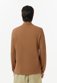 Brun långärmad polo med mjuk textur, som har en minimalistisk design och en liten krage, stylad med ljusa byxor.