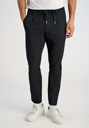 Uomo con pantaloni neri con coulisse, calzini bianchi e scarpe da ginnastica bianche, in piedi con una mano in tasca su sfondo uniforme.
