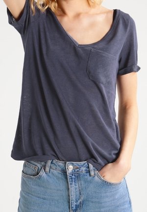 T-shirt en coton bleu marine à col V, à manches courtes et avec une poche avant. Porté ample sur un jean en denim bleu clair. Texture douce, design décontracté.