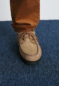 Bruna läderskors av båtmodell med rund tå, snörning och sömdetaljer; bärs med bruna kordflöjtsbyxor på en blå matta.