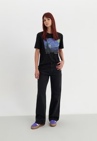 T-shirt noir avec un graphique de scène nocturne bleu et jaune, associé à un jean large noir et des baskets violettes avec des semelles brunes.