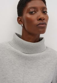 Grauer Rollkragenpullover mit geripptem Kragen und weicher Textur. Modell trägt einen kleinen silbernen Ohrring, der über einen dezenten Glanz und Designdetails verfügt.