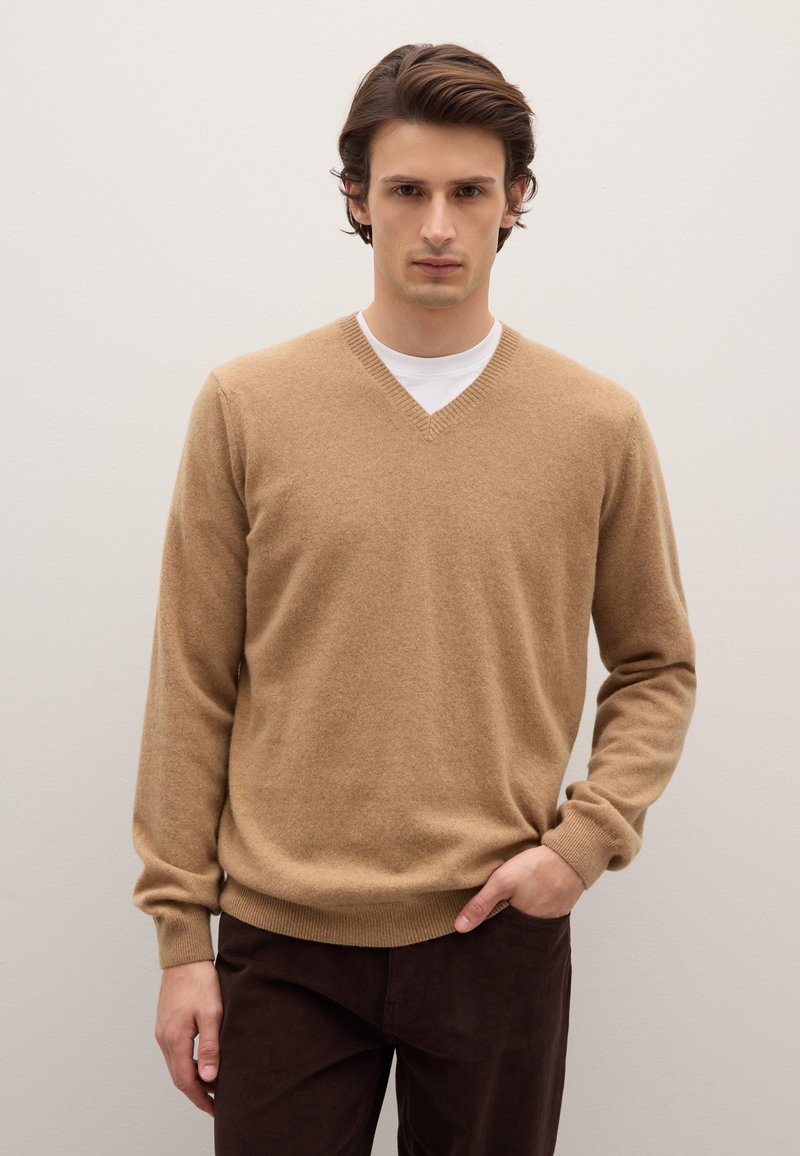 Hellbrauner Wollpullover mit V-Ausschnitt, gerippten Bündchen und Saum. Trägt man über einem weißen T-Shirt, kombiniert mit dunkelbraunen Hosen. Glatte Textur.