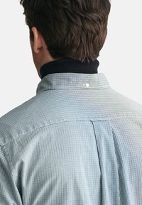Karierte Button-Down-Bluse mit einem dunklen gerippten Kragen, leichtem Stoff, Rückenschlitz und einem kleinen weißen Knopf an der Basis des Kragens.