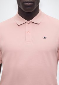 Homme portant un polo rose clair avec deux boutons fermés et un petit logo ovale sur le côté gauche de la poitrine.