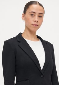 Svart kavaj med en texturerad tyg, slim fit, slagkragar och en enkel knappstängning. Bärs över en vit topp med subtila detaljer.