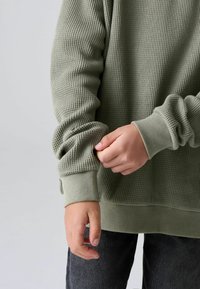 Felpa di colore verde oliva con texture a waffle e polsini a costine. Una mano aggiusta il polsino mentre sotto si vedono dei jeans scuri a contrasto.