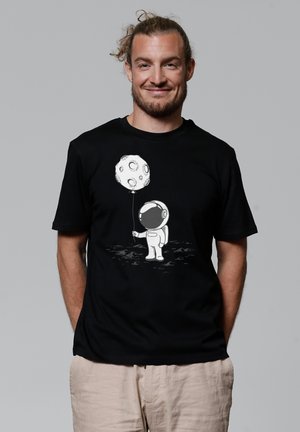 Zwarte T-shirt met een afbeelding van een witte, cartoonachtige astronaut die een maanvormige ballon vasthoudt tegen een achtergrond van een maanoppervlak.
