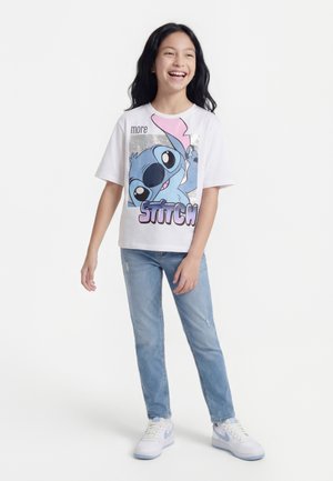 Ragazza sorridente con lunghi capelli scuri che indossa una maglietta bianca con la grafica di Stitch, jeans azzurri e scarpe da ginnastica bianche, in piedi su sfondo bianco.