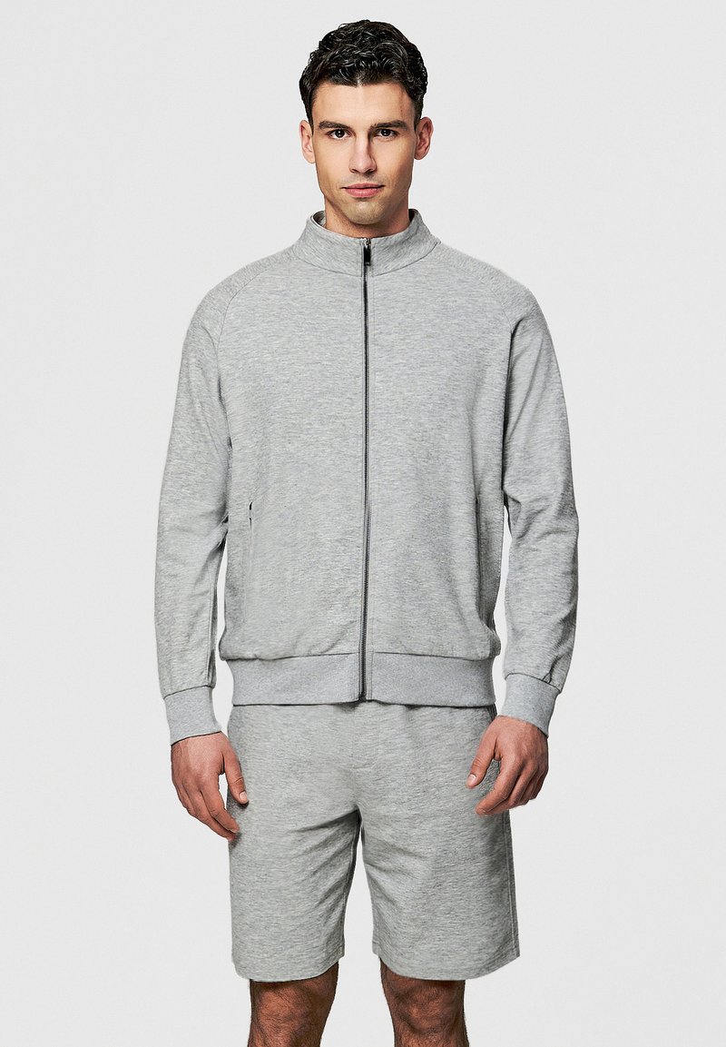 Młody mężczyzna ubrany w jasnoszary sweter na zamek i pasujące szorty, stojący na jednolitym tle.