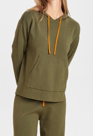 Femme portant un sweat à capuche vert olive avec des cordons orange et un pantalon assorti sur fond uni.