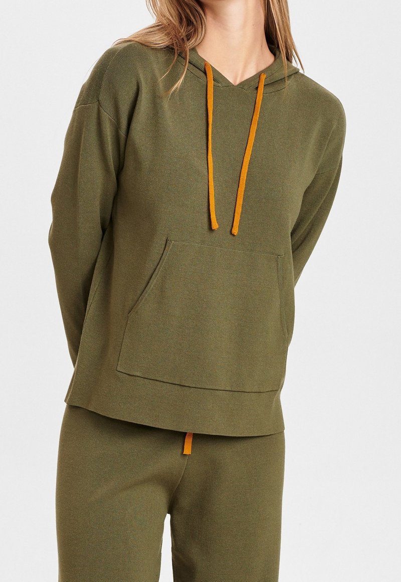 Femme portant un sweat à capuche vert olive avec des cordons orange et un pantalon assorti sur fond uni.