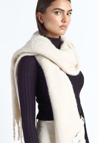 Kobieta w czarnej, prążkowanej bluzce z długim rękawem i grubej, miękkiej białej chuście narzuconej na ramiona na tle jednolitego białego tła.