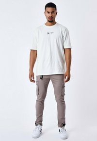 Camiseta de color beige claro con logotipo negro, combinada con pantalones cargo gris claro que tienen bolsillos laterales. Lleva zapatillas blancas.