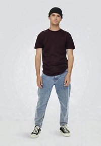Giovane uomo che indossa un berretto nero, una maglietta nera, jeans azzurri chiari e sneakers nere con lacci bianchi, in piedi davanti a uno sfondo chiaro e semplice.