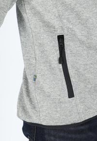 Giacca in maglia grigia con una tasca con zip nera e una piccola accentuazione blu e gialla. Tessuto morbido con una vestibilità aderente in vita.